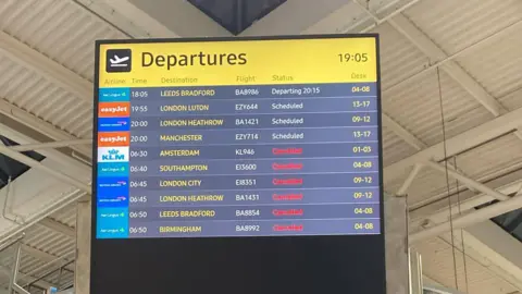 An elevated screen reads 'departures' in black font and a yellow background. Below that is a list of destinations with their flight status beside them. There are 10 destinations listed, six of them are cancelled. The other four haver not been cancelled. A white steel roof can be seen above the board.