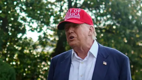 Donald Trump wears a red hat with white lettering reading Gulf of America wearing a white shirt and blue suit with no tie.