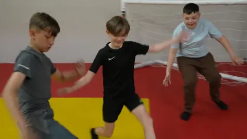 Three young boys playing football together on an indoor pitch. One is wearing grey trousers and a t shirt and has the ball, another is wearing all black and is trying to get the ball off him, and the other one is in goal and preparing for a shot.