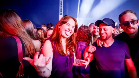 Fringe by the Sea A crowd at a gig, basked in red light. A woman and two men are in the foreground - the woman has long auburn hair, a purple vest top and is sticking her tongue our while making a 'devil horns' gesture with both hands. One of the men, wearing a blue cap smiles at her, while the other man, wearing sunglasses has his arm around him.