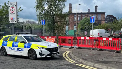 A police car parked in front of the school. The roof has been worn away by the fire. There is a red plastic fence in the foreground, parked behind a police patrol car.