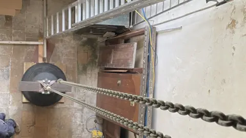 Shaun Whitmore/BBC A church bell, viewed from above, on a floor, with metal chains attached to it and leading upwards towards the camera 