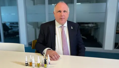 Alastair Richards is looking at the camera, wearing a suit and the tie is sitting on a table, sitting on a table with seven nose spray tanner in front of him