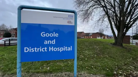 A photo of a blue and white sign that reads "Goole and District Hospital". There is grass and a tree in the background. 