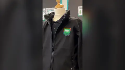 A black fleece with a logo saying 'AMA' on it on top of a black polo shirt worn by a mannequin