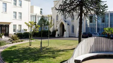 The image shows the exterior of a large cream-coloured building with a mix of traditional and modern architecture. There is an arched entrance in the centre, glass-fronted sections on the right, and a landscaped garden in front with a curved wooden bench, small trees, and a tall spiky tree. Several cars are parked near the glass section.
