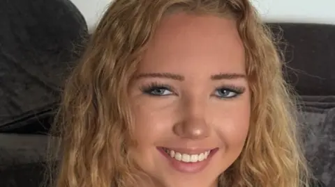 Family photo A young girl with curly blonde hair, she is wearing mascara and has long eyelashes. She is smiling at the camera 