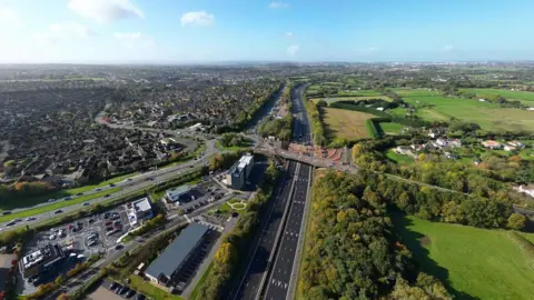 Andy Roberts/BBC A drone image shpws the M4 motorway close to Bristol with no traffic in one carriageway due to roadworks. Also visible is the Badminton Road Bridge where the work is taking place, with vehicles and large amounts of earth piled on the road surface. It is a sunny day with blue sky and the city of Bristol and surrounding countryside is also visible