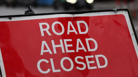 A red road sign reads 'ROAD AHEAD CLOSED'