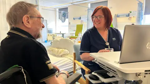 NHS Grampian A man with a beard and glasses in a black T-Shirt talks to a nurse in a blue uniform sitting at a keyboard