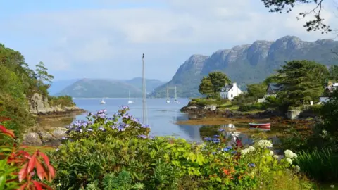 Debbie Neilson House, yachts in the bay and flowering plants in the foreground in Plockton