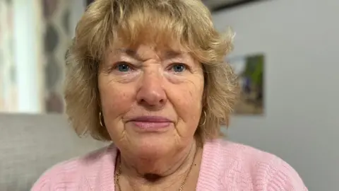 BBC Joyce Gifford with blue eyes and golden-coloured, short, wavy hair with a fringe. She is wearing a baby pink wool jumper with a thin, gold necklace and thin, gold, hoop earrings.