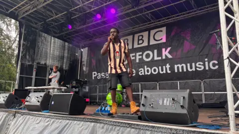 Ollie Ward/BBC A man sings into a microphone on a small music festival stage. He has dark hair and is wearing an orange and black striped football shirt, black shorts and brown Timberland-style boots. There is another man at one end of the stage DJing.