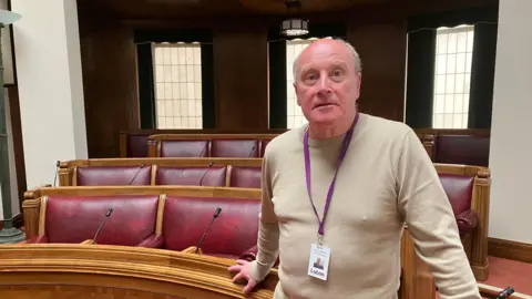 Amy Holmes/BBC Rob Roche, who is standing in the main chambers at Luton Borough Council. He is wearing a beige jumper and is stood in front of a number of red chairs.