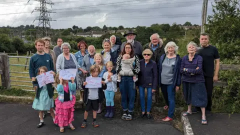 A large group of residents consisting of both adults and children. They are standing in front of the gate that leads into the field where the proposed facility would have been built. The children are holding hand drawn paper signs which say 'save nature'. The sky is grey and cloudy. In the distance you can see a large electricity power line from the neighbouring substation.