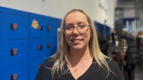 A woman with long blonde hair, glasses and a black top in front of rows of blue lockers