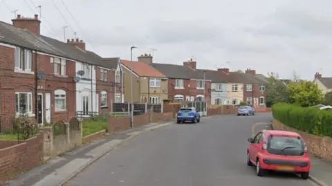 Google streetview image of a residential street with a few cars parked along it. It looks quiet and is a grey day. The houses are terraces and mostly unpainted brick with a few white and beige ones.
