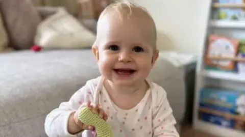 Family handout Baby Olivia holding a toy up to the camera while smiling directly into the lens.