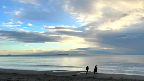 MarkieB The silhouettes of two people can be seen walking on a beach in the shadow of the sun obscured behind some light clouds. The water is calm with just a few wave ripples on the surface.