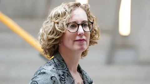 Lorna Slater, who has blonde hair and dark-rimmed glasses, in front of a stone staircase. She is wearing a grey jacket over a red top and turned side on to the camera. 