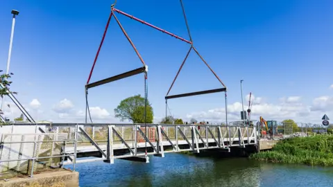 East Riding of Yorkshire Council A crane prepares to lift a 20-tonne section of Weel Bridge which spans the River Hull. The grey metal structure is set against a blue sky. Construction vehicles are visible in the distance.