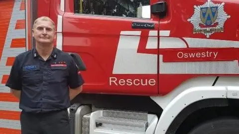 John Davies is standing in front of a red fire engine. He has his hands behind his back and is wearing a navy blue shirt and black trousers. The shirt has a Shropshire Fire and Rescue Service badge on the breast.