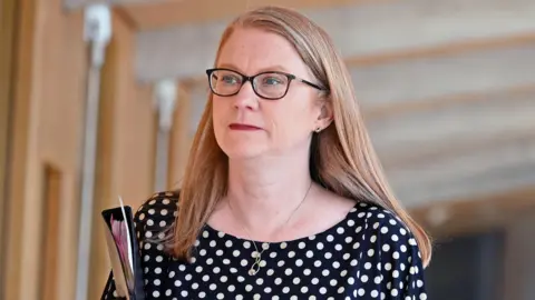 Getty Images Shirley-Anne Somerville - a a woman with glasses, shoulder length auburn and wearing a black dress with white polka dots - walks in a corridor at Holyrood while carrying a folder