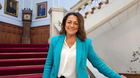 A woman standing on a staircase wearing a bright blue suit jacket and white shirt. She is smiling and looking directly at the camera.