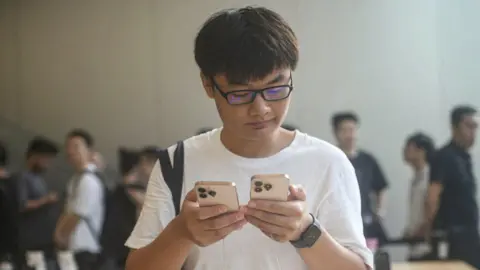 Getty Images Um jovem vestindo uma camiseta branca é mostrado em uma loja da Apple em Hangzhou, China, segurando um modelo do iPhone 16 em cada mão, com uma expressão pensativa no rosto.