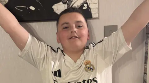 A boy with dark hair and a white football shirt on with both arms raised in the air