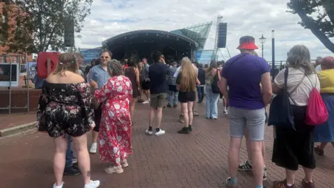 Several people are gathered in a square with a stage in the distance.