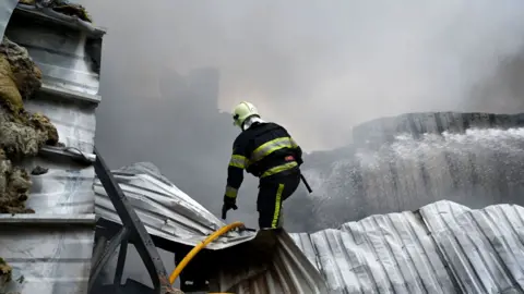 AFP via Getty Images A Ukrainian firefighter climbs the metal roof of a collapsed building to extinguish a fire at the site of a food warehouse following a Russian missile strike in Kyiv early on 25 October 2025