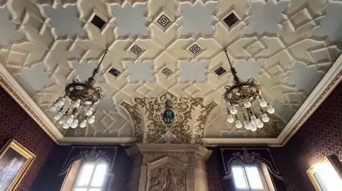 Sheffield City Council An ornate ceiling, carved and painted in cream and pale blue. Two large chandeliers hang down over tall windows. There is also an engraved tablet.