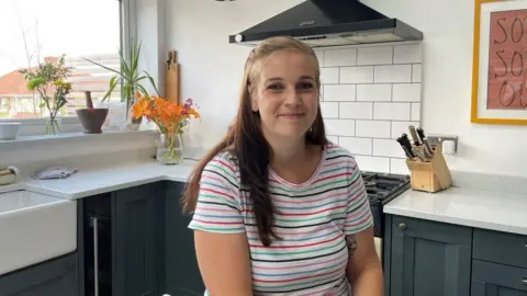 Lauren Peters sitting in her kitchen wearing a colourful striped t-shirt. The kitchen is light modern with white speckled countertops and dark grey cupboards. There are two vases of flours and a plant sitting on the windowsill. 