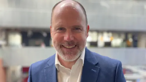 John MacKay, a bald man with a short, greying beard, is standing in BBC Scotland's Pacific Quay building. He is wearing a blue suit jacket with a white shirt, open at the neck. 