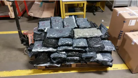 Stacks of vacuum-sealed rectangular packs of cannabis seized by police rest on a loading trolley in a warehouse.