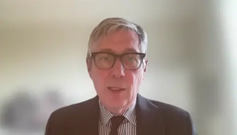 Eljamel Inquiry A man with grey hair and glasses, wearing a dark blue suit, a striped shirt and dark tie, speaks on a virtual call
