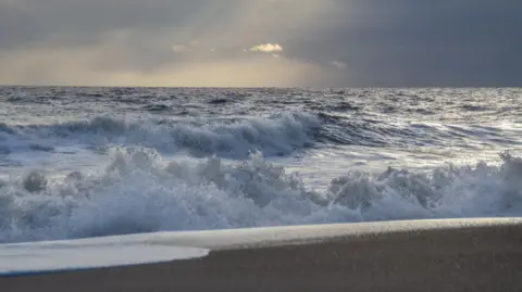 Big waves crash on to an empty stretch of sandy beach under a grey sky with rays of sunshine breaking through the clouds. 