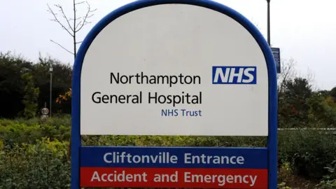 PA Media A white and blue sign next to bushes at the hospital. It says Northampton General Hospital NHS Trust, then below: Cliftonville Entrance, and in a red strip below that: Accident and Emergency.