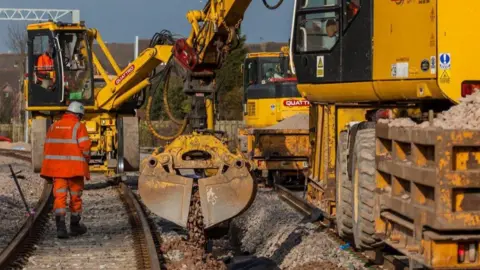 Network Rail A person wearing Hi-Vis and a helmet on a railway line with a yellow bit of machinery to the right of the image