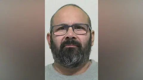 Mugshot of Hussain. He has balding short dark hair and a thick black and grey beard. He is wearing glasses and a grey t-shirt.