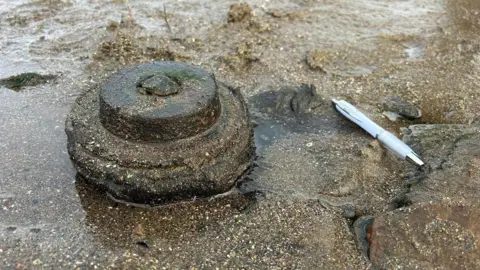 Whitehaven Coastguard The item found on the beach in Maryport, next to a pen, for scale. The item appears to be made of round sections progressively getting smaller towards the top. The item is a brown colour very close to the colour of the sand around it.