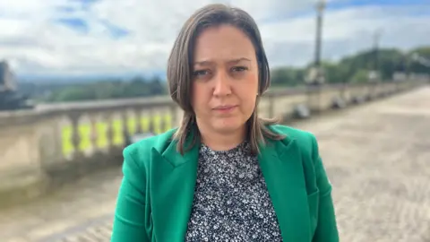 Deborah looking at the camera. She has shoulder length brown hair, a green blazer and a navy and white patterned top.