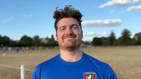 Elliot Deady/BBC A man - Joe Beastall - smiles straight at the camera while standing on a rugby pitch. Several players are behind him in the distance.