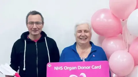 NHS Blood and Transplant A man and woman stand smiling at the camera next to some pink balloons. The woman holds a pink sign saying 'NHS Organ Donor Card' on it
