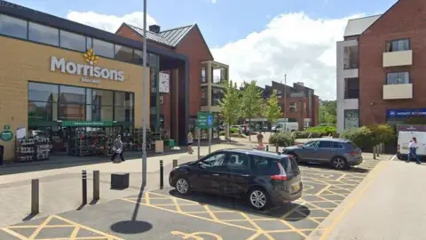 The outside of a Morrisons supermarket which has yellow bricks and a large glass entryway. There are disabled car parking spaces separated by yellow lines, with cars parked in them