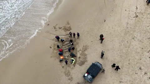 An aerial view of people gathered around the outline of part of the ship in the sand. There is a car parked nearby and some whitewash from the waves can be seen to the left.