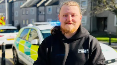 Kieran McPherson, a man with red hair tried back and a beard, and wearing a black hooded top, in front of an ambulance vehicle.