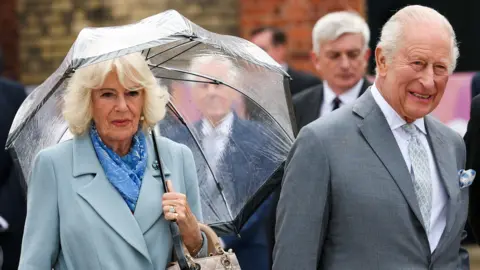 Reuters The Queen is on the left of the picture, she is wearing a light blue single breasted coat with a blue shawl. She is carrying a see-through umbrella. The King is on the right. He is wearing a grey suit with a pale lilac shirt. His tie is a pattern of pale grey and lilac. 