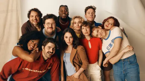Sky UK Posed picture of SNL UK cast smiling for the camera. They are standing against a beige backdrop. 